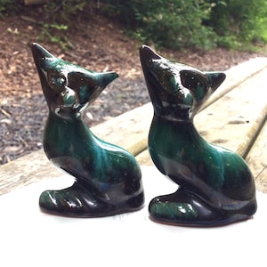 May include: Two ceramic cat figurines in a teal blue and black glaze. The cats are sitting upright with their heads tilted up.