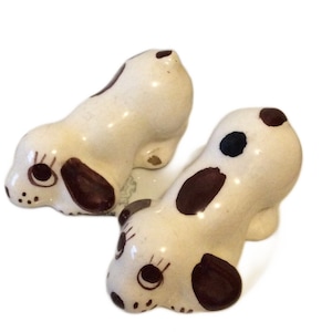 May include: Two ceramic dog figurines with brown spots and painted eyes. The dogs are white with brown markings and are in a playful pose.