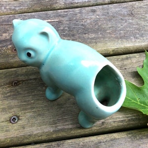 May include: A light blue ceramic planter shaped like a stylized animal, possibly a pig or a cat. The planter has a large opening on the back for planting.