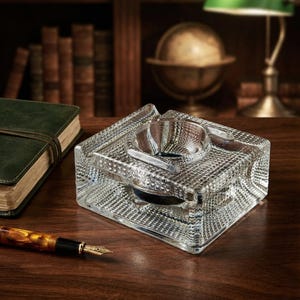 May include: A clear, rectangular glass inkwell featuring a textured, diamond-patterned exterior and a central circular reservoir. The inkwell has a thick, heavy base and a smooth, recessed top section designed to hold a writing instrument.