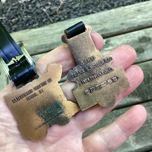 May include: Two bronze-toned metal luggage tags with black leather straps. One tag is embossed with CATERPILLAR TRACTOR CO. PEORIA ILL. The other tag is embossed with THE FRANK G. HOUGH CO. LIBERTYVILLE ILL. and includes a small logo with CHICAGO text.