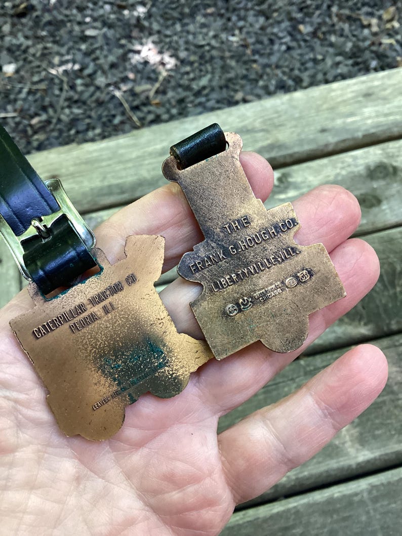 May include: Two bronze-toned metal luggage tags with black leather straps. One tag is embossed with CATERPILLAR TRACTOR CO. PEORIA ILL. The other tag is embossed with THE FRANK G. HOUGH CO. LIBERTYVILLE ILL. and includes a small logo with CHICAGO text.