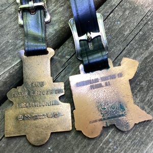 May include: Two bronze-toned metal luggage tags shaped like steam locomotives, each attached to a black leather strap with a silver-colored metal buckle. The left tag reads THE FRANK G. HOUGH CO. LIBERTYVILLE, ILL. The right tag reads CATERPILLAR TRACTOR CO. PEORIA, ILL.
