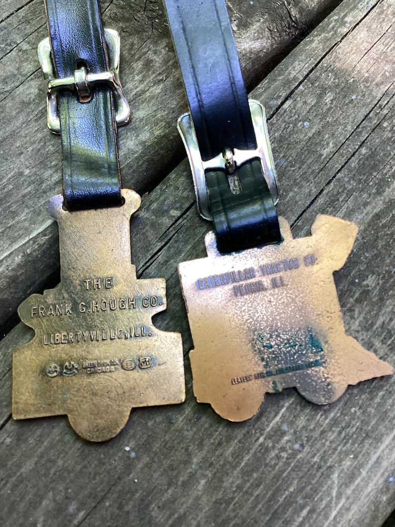 May include: Two bronze-toned metal luggage tags shaped like steam locomotives, each attached to a black leather strap with a silver-colored metal buckle. The left tag reads THE FRANK G. HOUGH CO. LIBERTYVILLE, ILL. The right tag reads CATERPILLAR TRACTOR CO. PEORIA, ILL.