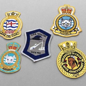 May include: Five embroidered patches featuring military-style emblems. One shield-shaped patch displays 'USS MIAMI SSN 755' with a palm tree. Four circular patches include designs with a crown, a wolf head, a red cross, and a profile of a person.