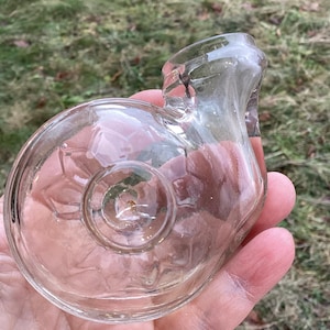 May include: A clear, vintage glass perfume bottle with a unique snail-shell design. The bottle has a rounded base with a central indentation and a curved neck. The glass is transparent, allowing a view of the contents.