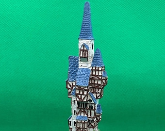 May include: A miniature figurine of a white and blue building with a pointed roof, perched atop a brown and green rocky cliff. The base of the figurine is signed "Mauritius 2014".