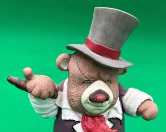 May include: A brown teddy bear figurine wearing a gray top hat with a red band, a white shirt, a purple jacket, a red and white patterned tie, and black and white checkered pants. The bear is standing on a brown wooden base.