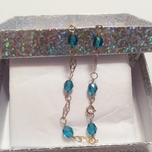 May include: A gold chain bracelet with teal blue glass beads. The bracelet is in a white gift box with a silver glitter pattern.