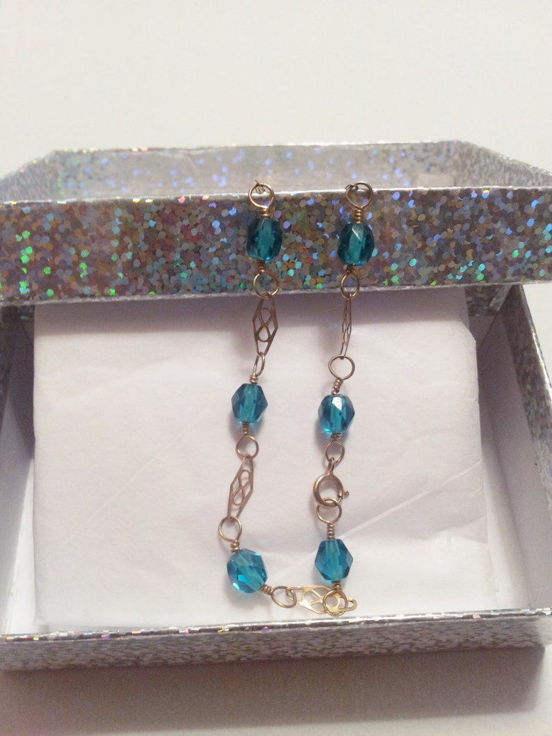 May include: A gold chain bracelet with teal blue glass beads. The bracelet is in a white gift box with a silver glitter pattern.