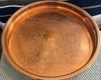 May include: A round, copper-colored serving tray with an ornate floral pattern. The tray has a decorative edge with a series of vertical slots and a rope-like border. The tray's surface is highly reflective, and the overall design is vintage.