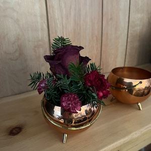 May include: A copper planter with three legs holds a bouquet of burgundy roses and carnations with green pine sprigs.