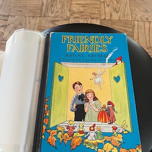 May include: A vintage hardcover book titled "Friendly Fairies" by Johnny Gruelle, author of the Raggedy Ann books. The cover features a blue background with a yellow and green window, a fairy flying, and a group of children watching a puppet show.