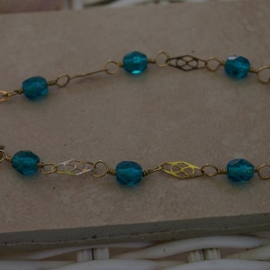 May include: A gold chain bracelet with teal blue glass beads. The chain has a delicate, intricate design.
