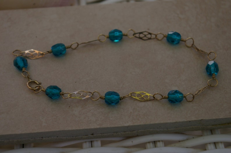 May include: A gold chain bracelet with teal blue glass beads. The chain has a delicate, intricate design.