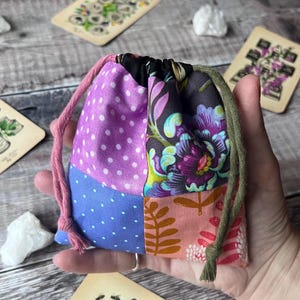 May include: A patchwork drawstring bag with pink, purple, blue, and floral patterned fabric. The bag has a pink drawstring and a green trim. Tarot cards and crystals are in the background.