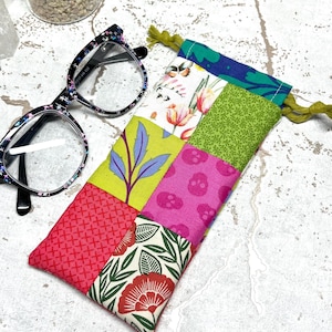 May include: A patchwork fabric eyeglass case with a drawstring closure. The case is made of different colourful fabrics with floral, leaf, and geometric patterns. The case is lying on a white surface.