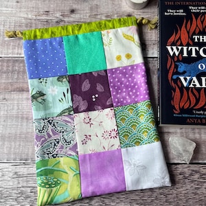 Peut inclure: Un sac à cordon patchwork avec une bordure verte et un cordon doré. Le sac est composé de divers carrés de tissu dans les tons de violet, vert et blanc, avec des motifs tels que des pois, des papillons et des motifs floraux. Un livre intitulé "The Witch of Val" est à côté du sac.