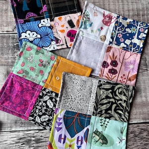 May include: A selection of handmade patchwork coasters. Each coaster showcases a unique blend of fabric patterns, such as mushrooms, florals, and geometric shapes. The colour scheme includes pink, blue, green, and black. These square coasters appear to be made of fabric.