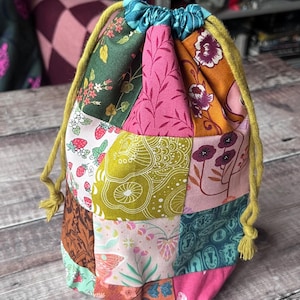 Op de afbeelding: Een patchwork-trekkoordtas met verschillende stoffen met bloemenpatronen in roze, groen, oranje en bruin. De tas heeft een gele trekkoordsluiting en staat op een houten oppervlak.