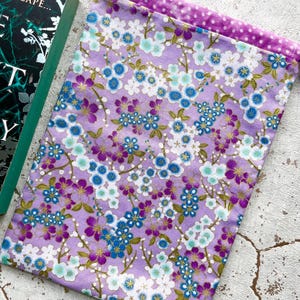 Peut inclure: Une pochette de livre en tissu lavande avec un motif floral de fleurs blanches, bleues et violettes. Le bord supérieur est doublé d'un tissu à pois violets et possède une fermeture à cordon. Un livre est partiellement visible.
