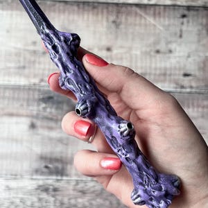 May include: A purple wand with a textured, branch-like design. The wand has silver accents and three circular details. The wand is held in a hand with red nail polish.