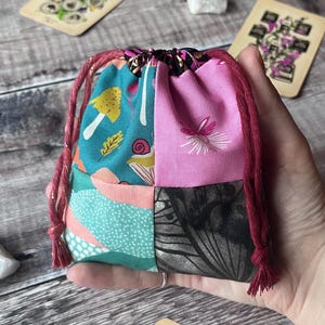 May include: A small, patchwork drawstring bag with a variety of colorful fabric patterns. The bag features teal, pink, and black panels with whimsical designs, including mushrooms, snails, and butterflies. The drawstring is a deep red with gold accents.