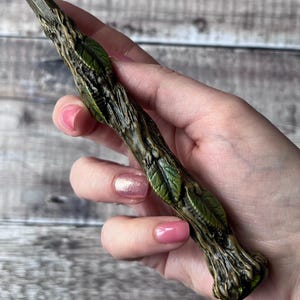 May include: A detailed wand with a textured, brown wooden handle and green leaf accents. The wand's tip is a darker shade of green. The wand is held in a hand with pink nail polish.