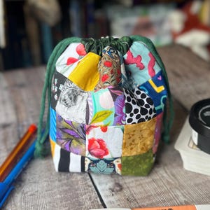 May include: A patchwork drawstring bag with a green drawstring. The bag is made from various fabric squares in different colours and patterns, including floral, polka dot, and abstract designs. Pens and highlighters are in the foreground.