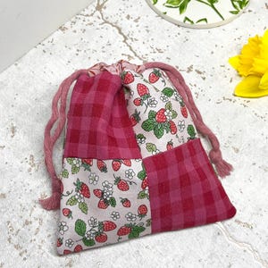 May include: A pink and white drawstring bag with a patchwork design featuring a strawberry print and pink gingham. The bag is made of fabric and has a drawstring closure.