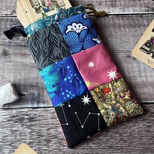 May include: A patchwork tarot card bag with a variety of patterned fabrics in shades of blue, pink, and black. The bag is filled with tarot cards, and the drawstring is green. The bag is on a wooden surface.