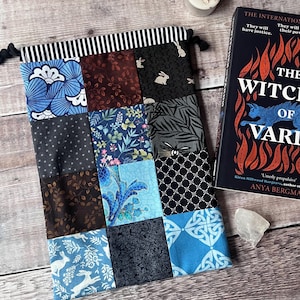May include: A patchwork book sleeve with a black and white striped top and a drawstring closure. The sleeve features various fabric patterns, including floral, geometric, and rabbit motifs. A book titled "The Witch of Varl" is next to the sleeve.