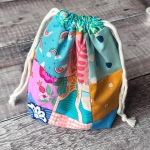 May include: A patchwork drawstring bag with a variety of colourful patterns, including rainbows, flowers, and abstract shapes. The bag is made of fabric and has a white drawstring closure. The bag is sitting on a wooden surface.