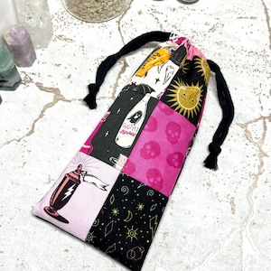 May include: A black drawstring bag with a patchwork design featuring pink, black and white fabric. The fabric has various illustrations including skulls, a ghost, a bottle, a sun and a crescent moon.