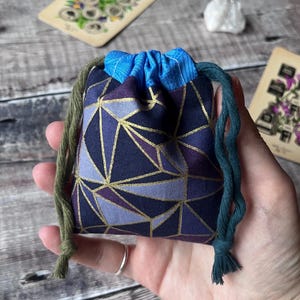 May include: A small drawstring pouch with a geometric pattern in shades of blue, purple, and gold. The pouch has a blue top and green and teal drawstrings. Tarot cards and a crystal are in the background.