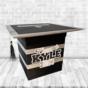 May include: Black graduation cap shaped card box with silver trim and a black tassel. The box has silver and black stripes and a sticker that says "Congrats Kyle 2024".