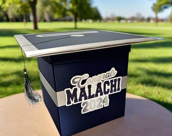 May include: A navy blue square graduation card box featuring a silver ribbon band and a graduation cap topper. The front displays the text "Congrats MALACHI 2024" in white and silver lettering. A black tassel hangs from the side of the cap topper.