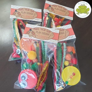 May include: Four clear plastic bags filled with craft supplies for making a play dough turkey. Each bag contains pipe cleaners, pom poms, googly eyes, and a small lid. The bags have a printable label that says "Make Your Own Play Dough Turkey".