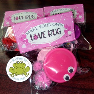 May include: Pink and white bag with a pink and white label that says "Make Your Own Love Bug". The bag contains a pink lid, two pink pom-poms, two googly eyes, and a green frog sticker that says "One Stop Mommy Shop".