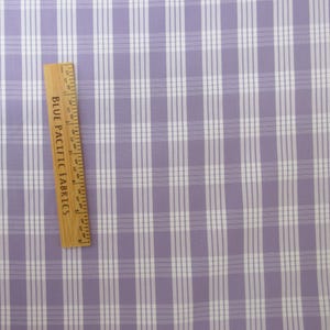 May include: A close-up of a fabric with a plaid pattern in lavender and white. A wooden ruler with the text "BLUE PACIFIC FABRICS" is placed diagonally across the fabric. The ruler measures up to 5 inches.