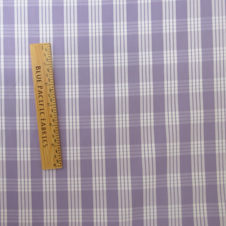 May include: A close-up of a fabric with a plaid pattern in lavender and white. A wooden ruler with the text "BLUE PACIFIC FABRICS" is placed diagonally across the fabric. The ruler measures up to 5 inches.