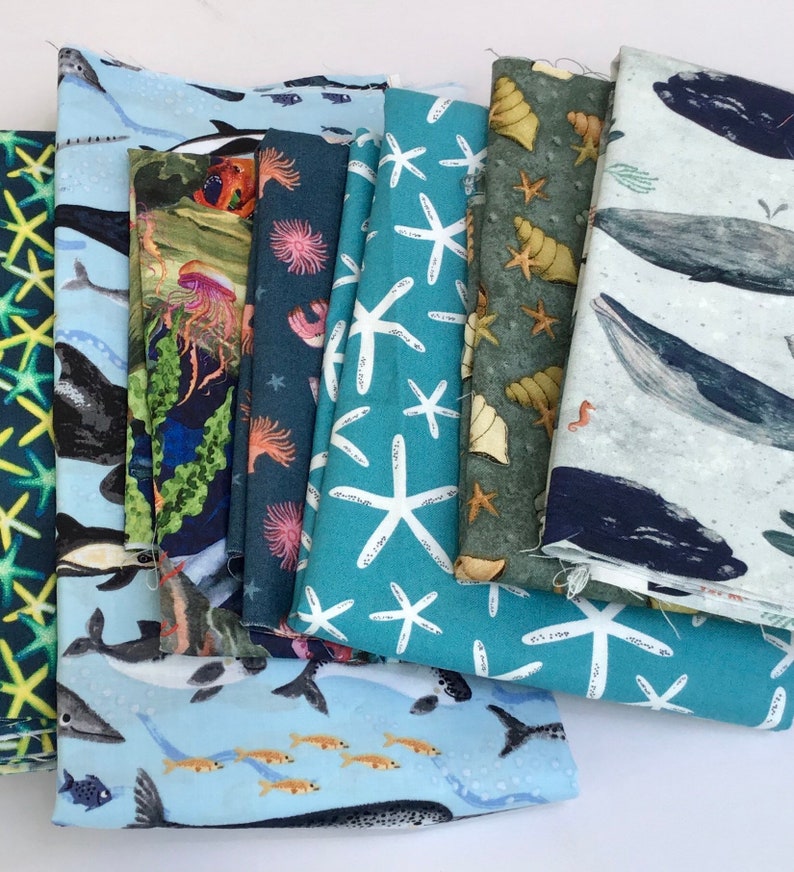 May include: A stack of eight fabric squares with different ocean-themed patterns. The patterns include starfish, seashells, whales, and coral reefs. The fabrics are in shades of blue, green, and white.