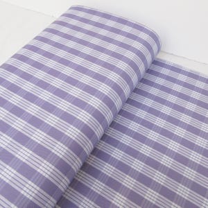 May include: A roll of fabric with a lavender and white plaid pattern. The fabric is rolled up, revealing the pattern of intersecting lines. The fabric is a light color and appears to be cotton or a similar material.