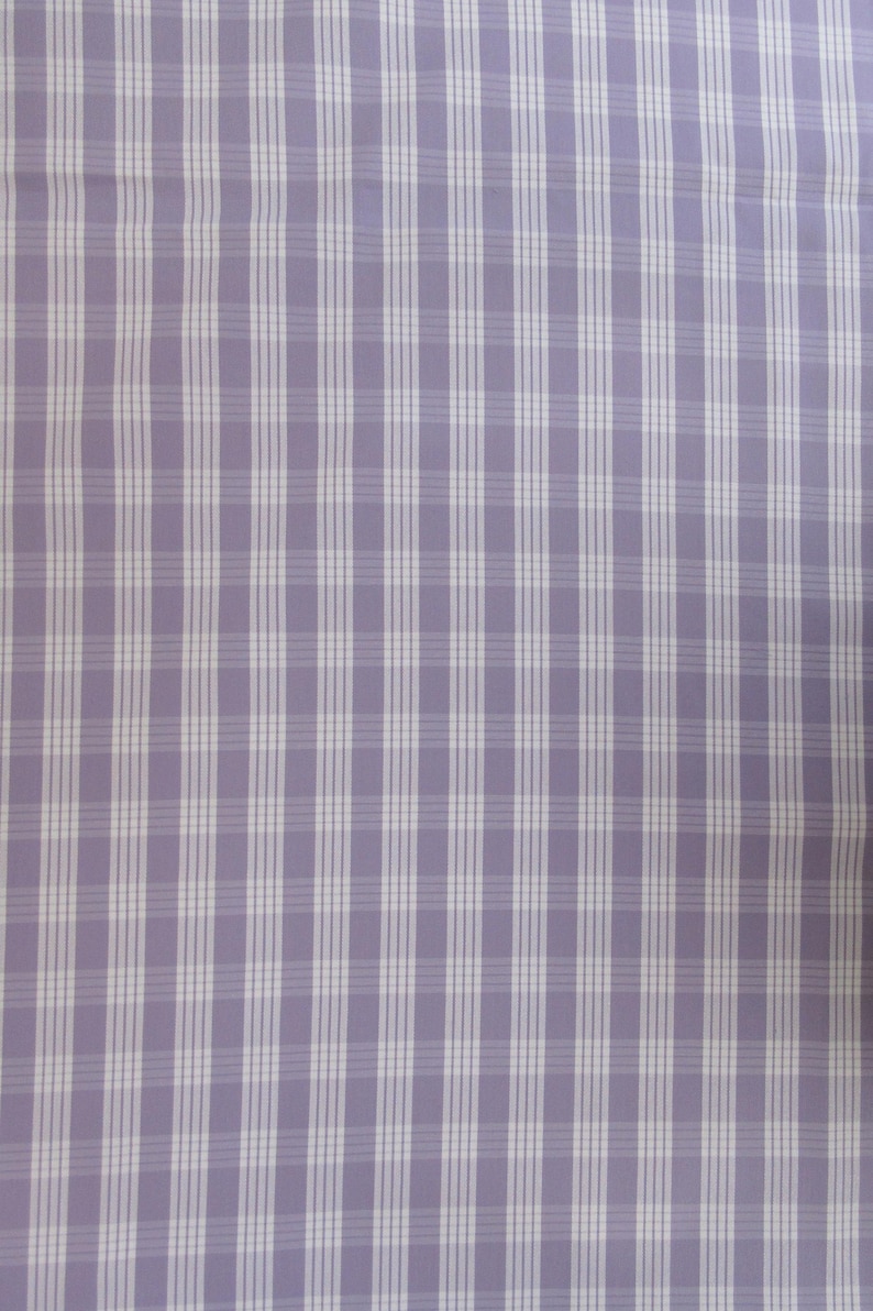 May include: A close-up of a fabric with a lilac background and a white plaid pattern. The plaid consists of vertical and horizontal lines, creating a grid-like design. The fabric appears to be smooth and possibly used for clothing or home decor.