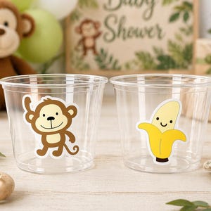 May include: Two clear plastic cups with cartoon stickers of a monkey and a banana. A plush monkey and a sign that says "Baby Shower" are in the background. Balloons and a wooden toy are also visible.