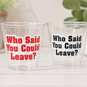 May include: Two clear plastic cups with the text "Who Said You Could Leave?" in red and black. The cups are on a white wooden surface, with greenery in the background.