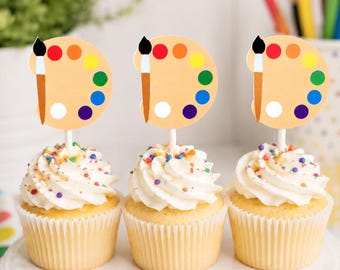 May include: Three cupcakes with white frosting and colourful sprinkles, each topped with a circular paper topper featuring a paintbrush illustration and a paint palette with red, orange, yellow, green, blue, purple, and white paint circles.