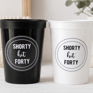May include: Two plastic cups, one black and one white, each with a circular sticker. The stickers read "SHORTY hit FORTY" in a black and white design. The cups are likely for a birthday celebration.