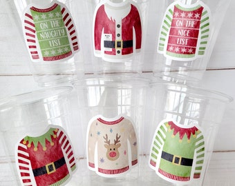 May include: Six clear plastic disposable cups, each featuring a sticker of a different holiday sweater design. The sweaters include a green one with text reading ON THE NAUGHTY LIST, a red Santa suit, a red one with ON THE NICE LIST, and elf and reindeer motifs.