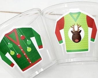 May include: Three clear plastic cups with festive Christmas sweater designs. Each cup features a different sweater design: one with ornaments, one with a reindeer, and one with a Christmas tree. The sweaters are in shades of green, red, and white.